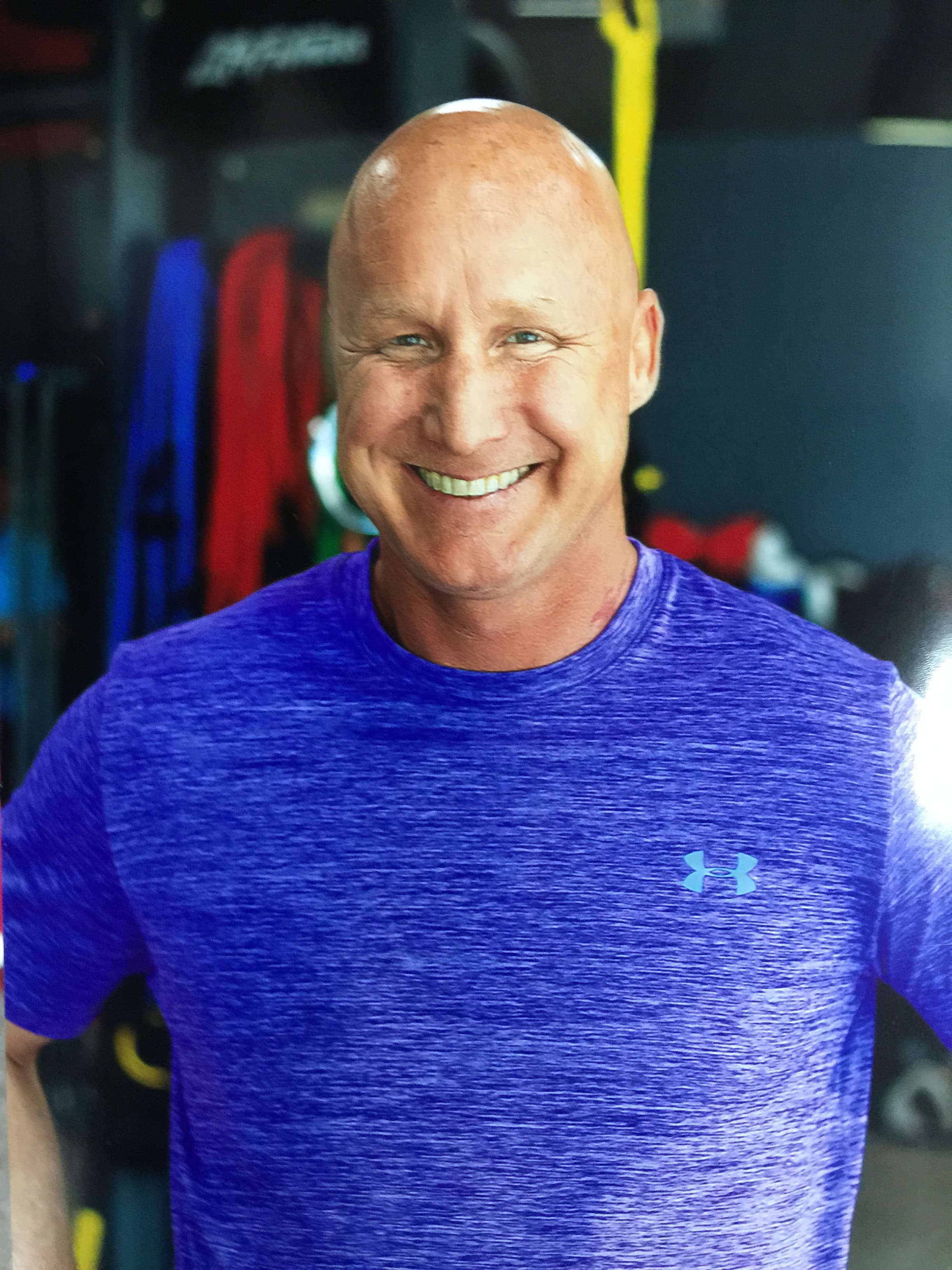 Smiling bald man in a purple athletic shirt posing in a gym with equipment.